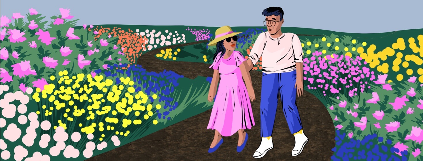A couple is walking together through a flowered path. A woman wears a hat and sunglasses.